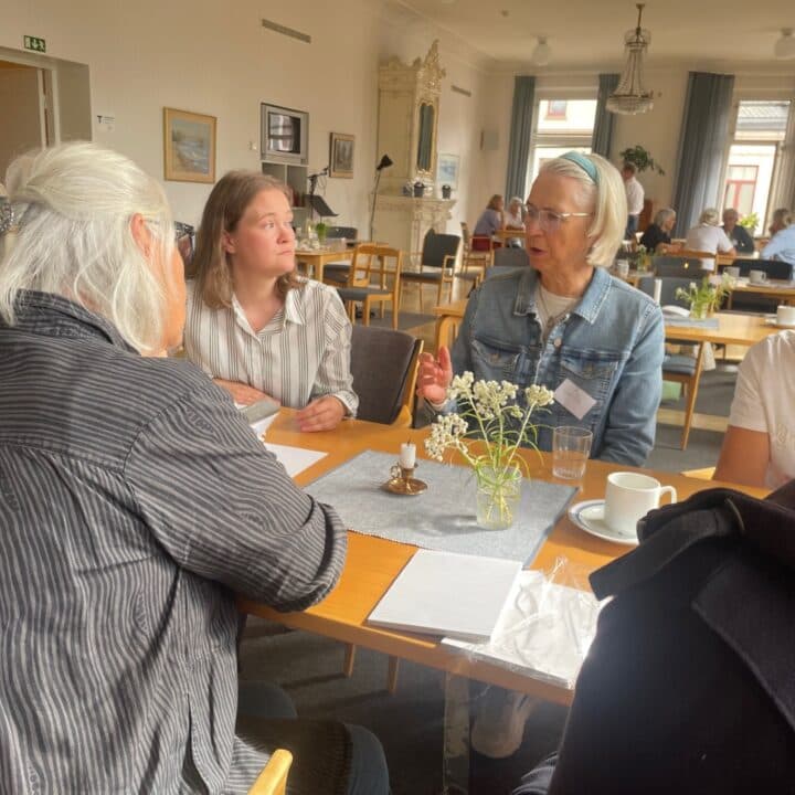 Fem personer sitter kring ett bord i en samlingssal och pratar/lyssnar aktivt på varandra.