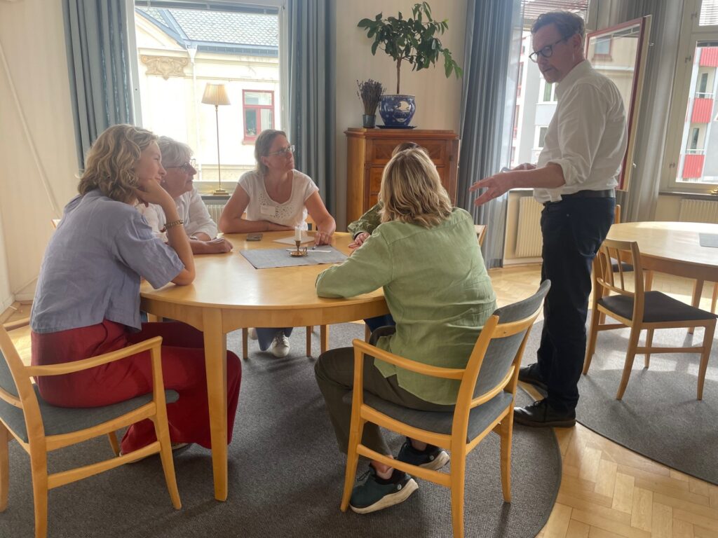 fyra kvinnor runt ett bord, som vänder uppmärksamheten mot en man som står intill bordet och gestikulerar.